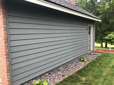 Side view of a gray house with a brick corner, white trim, a small door, and a landscaped area with rocks and green plants.