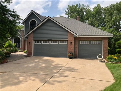 Gray house with brick accents, a three-car garage, arched windows, and a concrete driveway, surrounded by trees and landscaped areas.