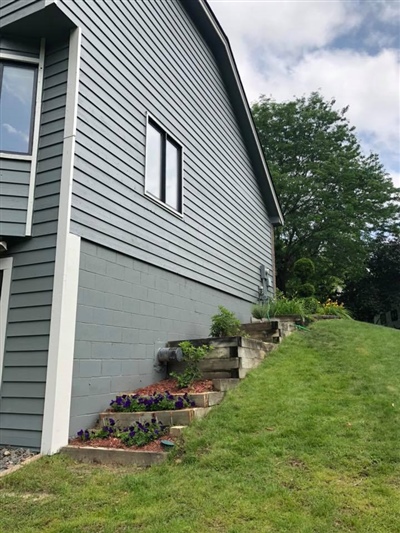 Side view of a gray house with a sloped lawn, brick steps, and flower planters leading up to a higher level, with trees in the background.