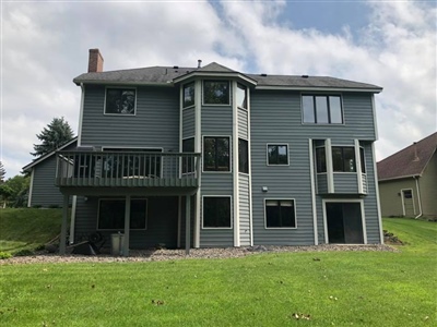 Back view of a large gray house with multiple windows, a balcony, and a sloped lawn extending to the house's foundation.