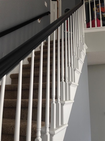 An indoor staircase with white spindles and a dark handrail, carpeted steps, and a gray wall, leading up to a second-floor railing.