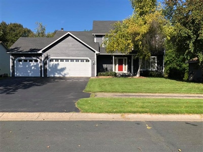 Front view of a gray house with a red front door, a two-car garage, a black driveway, and a well-kept lawn with trees.