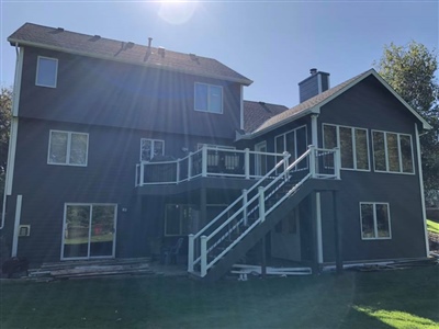 Back view of a gray house with multiple windows, a balcony, and an exterior staircase leading to an upper level, with sunlight streaming over the roof.