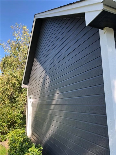 Side view of a gray house with white trim, a clear blue sky, and trees in the background.