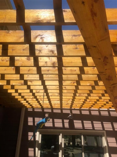 View of a wooden pergola casting shadows on a patio with glass doors, under a clear blue sky.