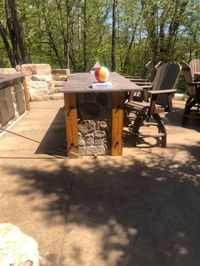 Outdoor bar with a stone base and wooden countertop, surrounded by high wooden stools, on a concrete patio with trees in the background.