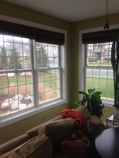 A cozy indoor sitting area with large windows featuring white frames and brown blinds, overlooking a green yard and a neighborhood.