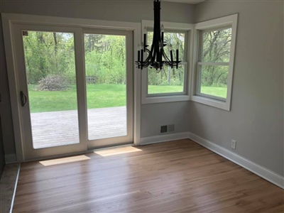 Dining area with light wood flooring, a black chandelier, large sliding glass doors leading to a patio, and two corner windows providing a view of the greenery outside.