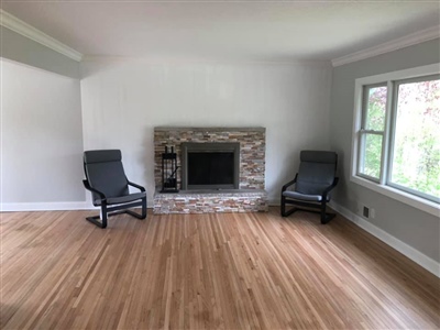 Living room with light wood flooring, a stone fireplace, two black chairs, white walls, and a window on the right side.