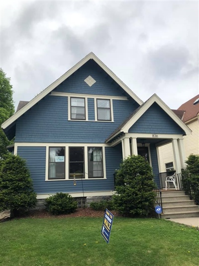 Two-story blue house with white trim, a front porch, and manicured shrubs, set on a green lawn under a cloudy sky.