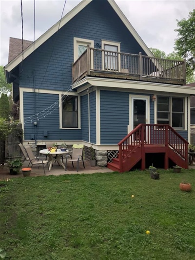 Back view of a blue house with white trim, a wooden deck, a balcony, and a green lawn with some outdoor furniture and potted plants.