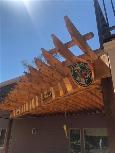 Wooden pergola with a decorative sign, casting shadows on the patio below, under a clear blue sky.