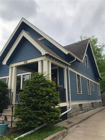 Side view of a blue house with white trim, a front porch, and green shrubs, set along a concrete path under a cloudy sky.