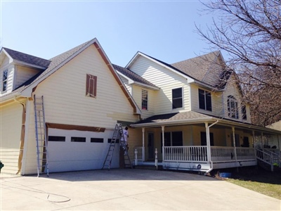 A large two-story house with light yellow siding, a spacious front porch, and a double garage, with a ladder propped against the side.