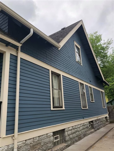 Side view of a blue house with white trim and a stone foundation, featuring several windows and set along a concrete path under a cloudy sky.