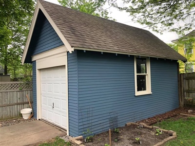 Small blue garage with white trim, a pitched roof, and a single window, set in a fenced yard with trees.