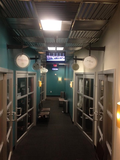 Dimly lit hallway with teal walls, glass doors on both sides, hanging signs, and a TV screen at the end displaying information.