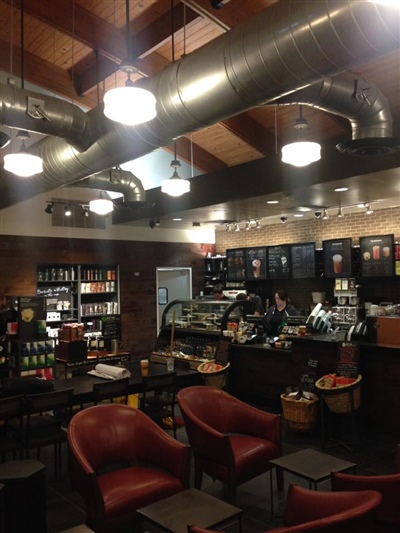 Cozy coffee shop with exposed ductwork, pendant lights, shelves stocked with products, a counter with baristas, and seating with red chairs and small tables.