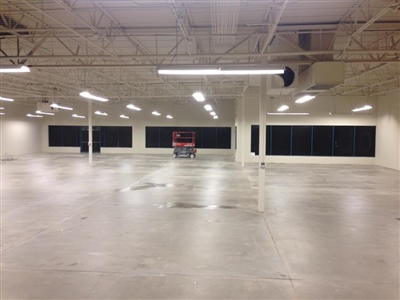 Large empty warehouse with white walls, exposed ceiling, and concrete floor, featuring large windows along one side and a scissor lift in the center.