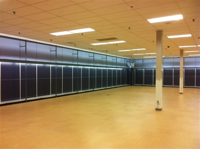 Empty commercial space with yellow flooring, white ceiling tiles, and a row of large glass-front refrigerators along one wall.