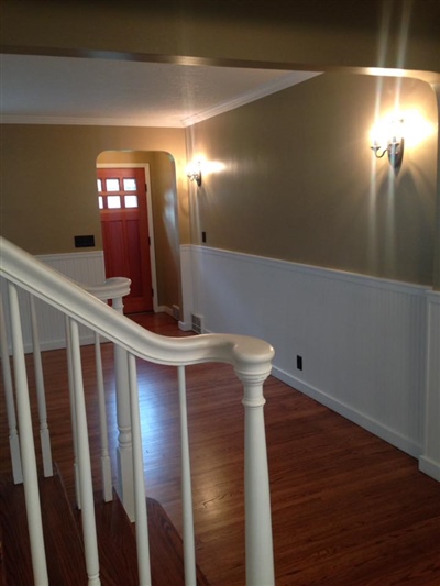 A hallway with wooden floors, beige walls, white wainscoting, and wall sconces, leading to a red front door.