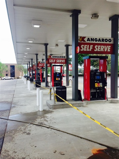 Gas station with multiple red and black fuel pumps under a white canopy, featuring "Kangaroo Self Serve" signage and yellow caution tape on the ground.