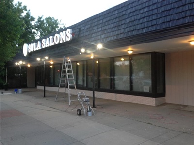 Exterior of a strip mall with a sign reading "Sola Salons," featuring large windows, a blue awning, and a ladder set up on the sidewalk.