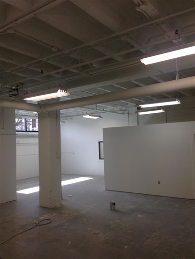 Empty industrial space with white walls, concrete floors, exposed ceiling beams, and fluorescent lighting.