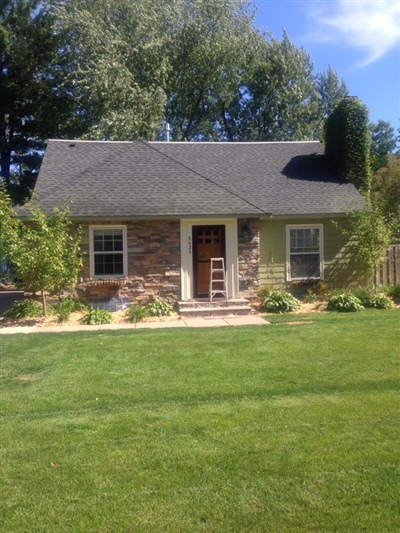 A small cottage-style house with a mix of stone and light green siding, a central front door, and a well-maintained lawn with surrounding greenery.