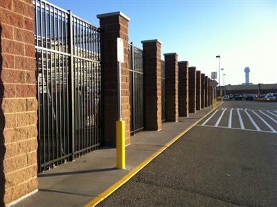 A fenced area with brick columns and black metal gates along a paved walkway, with a yellow bollard in the foreground.