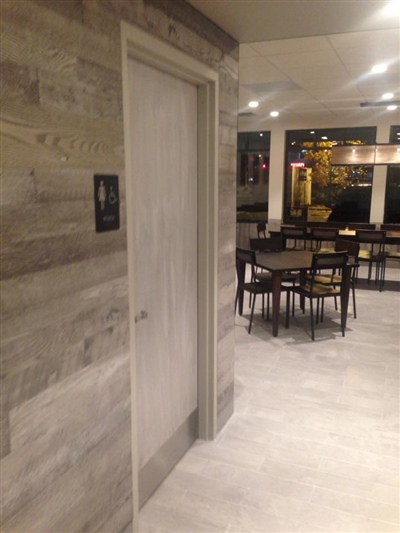 A modern interior with a light-colored wooden door on the left and a dining area with tables and chairs in the background.