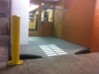 An entrance to a building with a ramp, yellow bollard, and glass doors, featuring a striped walkway and tan walls.