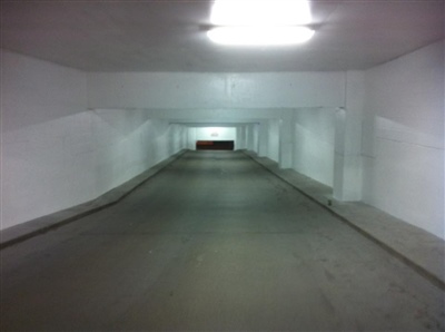 An empty, well-lit underground parking garage with white walls and a long, straight ramp leading to an exit.