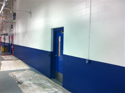 A hallway with white and blue walls, a blue door in the middle, and exposed pipes and cables on the ceiling.