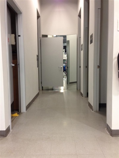 A corridor with gray doors on both sides, leading to an open door at the end, revealing a room with shelves.