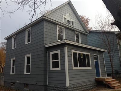 A two-story house with gray siding, white trim, and a blue front door, featuring a small porch and surrounded by bare trees.