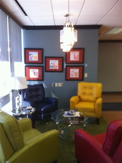 A cozy seating area with colorful chairs, a glass coffee table, and framed artwork on the wall, illuminated by a hanging light fixture.