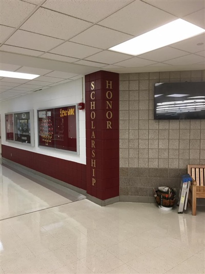 A school hallway with display cases, a maroon column with the words "Scholarship" and "Honor," and a tiled wall.