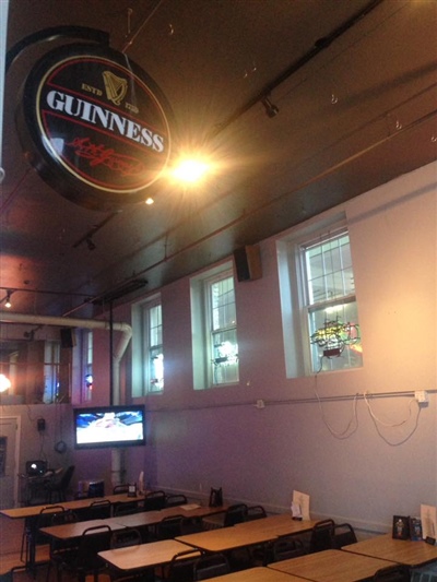 A pub interior with wooden tables, a TV on the wall, and a hanging Guinness sign, featuring windows on the side.