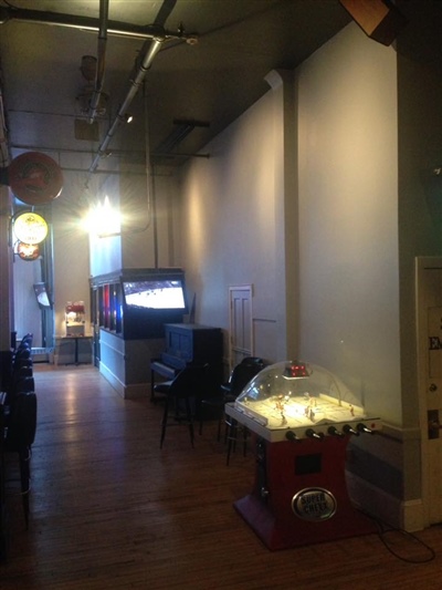 A pub interior with wooden floors, a TV on the wall, and a coin-operated game machine, featuring hanging lights and signs.