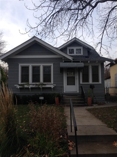 A small gray house with white trim, a front porch, and a central walkway leading to the front door, surrounded by a garden and bare trees.