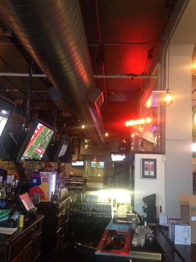 A pub interior featuring a bar counter, multiple TVs showing sports, exposed ductwork on the ceiling, and various bar equipment.