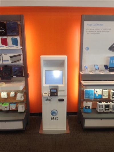 An AT&T store display with an orange background, featuring a self-service kiosk flanked by shelves of phone accessories and gadgets.