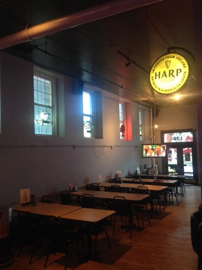 A pub interior with wooden tables and chairs, a Harp beer sign hanging from the ceiling, and windows along the wall.