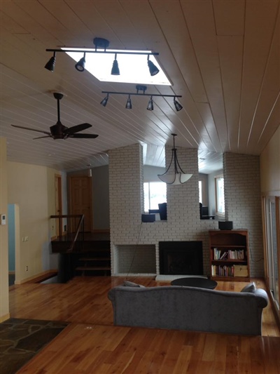 A modern living room with wooden floors, a skylight, track lighting, a ceiling fan, and a cozy seating area facing a fireplace and bookshelves.