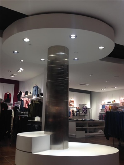 A modern retail store interior featuring a central column with circular, illuminated display platforms and shelves with various clothing items in the background.