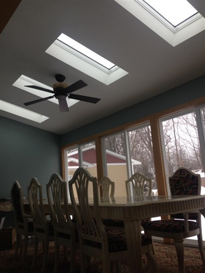 A dining room with a long table and chairs, featuring skylights, a ceiling fan, and large windows showing a snowy outdoor scene.