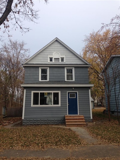 A two-story gray house with white trim and a blue front door, featuring a small porch and surrounded by autumn trees with fallen leaves.