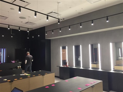 A modern retail store interior featuring illuminated mirrors, track lighting, black counters, and a person working in the background.