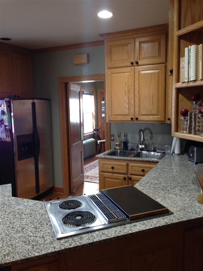 A cozy kitchen with wooden cabinets, granite countertops, a stainless steel refrigerator, and an electric cooktop, leading to a bright hallway.